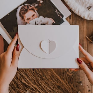 Può includere: Un invito di nozze bianco con un ritaglio a forma di cuore, tenuto in mano con smalto rosso. All'interno dell'invito è visibile una fotografia di una sposa. Lo sfondo è una superficie di legno con fiori secchi ed elementi decorativi.