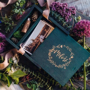 Puede incluir: Una caja de madera verde azulado con tapa, que contiene fotografías, una pequeña botella de madera y piñas. La tapa está decorada con una corona floral y la palabra "Miłość". La caja está rodeada de flores y vegetación.