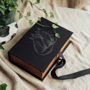 May include: A black book with a detailed illustration of two foxes on the cover. The book has a wooden spine and a black ribbon. Green ivy and a decorative stone are in the background.