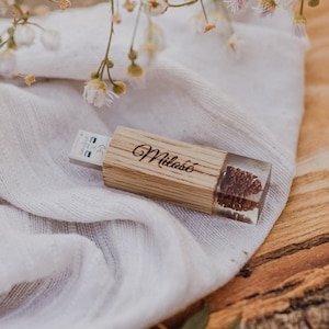 May include: A wooden USB flash drive with the word "Miłość" engraved on it. The end of the drive is encased in clear resin with a pinecone inside. The drive rests on a white cloth with floral accents.
