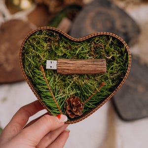 May include: A wooden USB flash drive rests in a heart-shaped box filled with green moss and pine needles. A small pine cone sits at the bottom. The box is held in a person's hand.