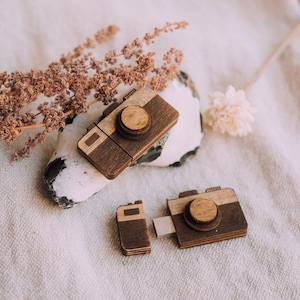 Op de afbeelding: Houten USB-sticks in de vorm van camera's in verschillende maten. De sticks zijn gemaakt van donker- en lichtbruin hout, met een cirkelvormig lensdetail. Ze zijn gerangschikt op een witte steen en een textuurrijke, gebroken witte stoffen achtergrond.