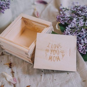 Może przedstawiać: Małe, otwarte drewniane pudełko z pokrywką. Na pokrywce wygrawerowano słowa "I LOVE YOU MOM" ozdobną czcionką, otoczone elementami kwiatowymi. W pobliżu ułożono fioletowe kwiaty bzu.