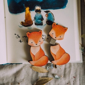 May include: Two orange and white fox-shaped magnets are displayed on an open book, alongside a watercolor illustration. The magnets are positioned near the text "So much to..." on the page. The scene is set on a textured, neutral-toned surface.