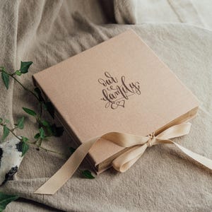 Può includere: Un album fotografico quadrato, marrone chiaro, con la scritta "our family" in corsivo sulla copertina. Un nastro beige è legato attorno all'album. L'album è su una superficie di stoffa beige, con piante verdi sullo sfondo.