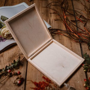 May include: An open, light-colored wooden box with a hinged lid, resting on a wooden surface. The box is empty, revealing its interior. The scene includes a magazine, flowers, and foliage.
