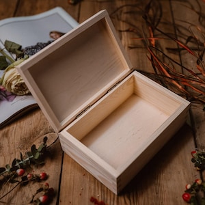 Könnte beinhalten: Eine helle Holzkiste mit offenem Deckel, die auf einer Holzoberfläche steht. Die Kiste ist leer und zeigt ihr Inneres. Ein Magazin, eine Rose und Zweige mit Grünzeug sind um die Kiste herum angeordnet.