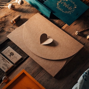 Può includere: Un tavolo rustico in legno espone articoli di cancelleria. Una busta di carta marrone con un ritaglio a forma di cuore è il punto focale. Altri oggetti includono una carta verde acqua con scritte dorate, timbri in legno e un piccolo vassoio arancione.