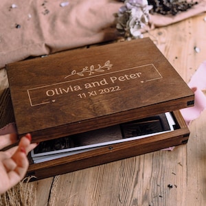 Könnte beinhalten: Eine Holz-Fotobox mit den Namen "Olivia und Peter" und dem Datum "11 XI 2022" auf dem Deckel graviert. Die Box ist geöffnet und zeigt Fotos im Inneren. Ein rosa Band und Trockenblumen sind ebenfalls zu sehen.