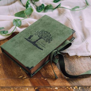 Puede incluir: Un diario verde con una ilustración de un árbol y un conejo en la portada, atado con una cinta verde oscuro. El diario está sobre una superficie de madera con una tela blanca y vegetación en el fondo.