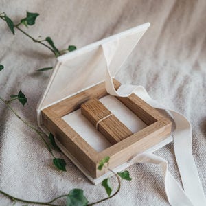 Può includere: Una scatola aperta, bianco sporco, con una cornice in legno e un oggetto rettangolare in legno all'interno. Un nastro bianco è attaccato alla scatola. La scatola è su una superficie di colore neutro, con verde e un candelabro sullo sfondo.