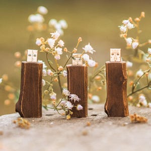 May include: Three wooden USB flash drives with a natural wood grain finish are displayed on a weathered surface. Each drive is mounted on a small, dark brown wooden base. Delicate white flowers and greenery surround the objects.