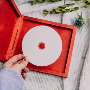 May include: A red wooden box with a white CD inside. The CD has a red center. The box is open, revealing the CD. The background includes greenery and a white cloth.