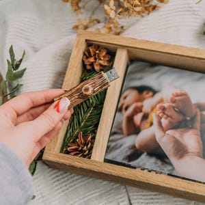 May include: A wooden USB drive with a natural wood grain design is held near a wooden photo frame. The frame contains a photograph of a baby's feet. The frame is decorated with pinecones and greenery.