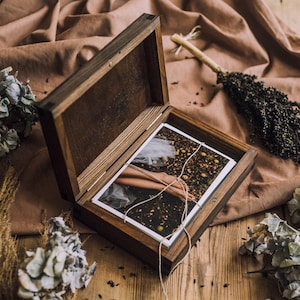 Könnte beinhalten: Eine dunkle Holzkiste mit einem Klappdeckel, die geöffnet ist, um ein Foto im Inneren zu zeigen. Die Kiste ist mit Bindfaden gebunden und liegt auf einer Holzoberfläche. Getrocknete Blumen und Blätter umgeben die Kiste.