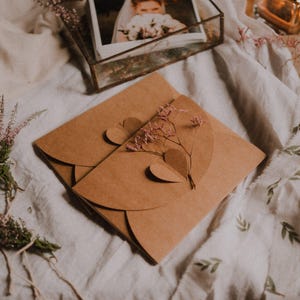 Può includere: Un invito di carta marrone con un design a forma di cuore e fiori secchi. L'invito è su un panno bianco con una foto in una scatola di vetro e piccoli bouquet di fiori viola.
