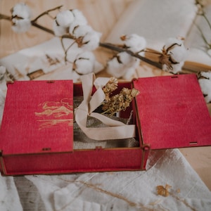 Könnte beinhalten: Eine rote Holzkiste mit offenem Deckel, die ein beiges Band und getrocknete Blumen zeigt. Die Kiste hat ein Landschaftsdesign auf dem Deckel. Baumwollzweige und ein Buch sind im Hintergrund.
