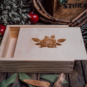 Può includere: Una scatola di legno color chiaro con coperchio scorrevole, con un'incisione dettagliata di una rosa. La scatola è aperta, rivelando un interno vuoto. Lo sfondo include elementi decorativi.