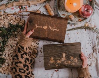 May include: Two wooden boxes with engraved designs. The top box has the names "Anna" and "Mateusz" with a heart symbol. The bottom box features an illustration of people with cameras. The boxes are dark brown and held by a person wearing a brown sweater.