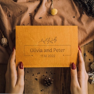 Puede incluir: Placa de madera grabada con los nombres "Olivia y Peter" y la fecha "11 XI 2022" en un marco rectangular. La placa tiene un diseño floral. El fondo incluye flores secas y una tela marrón.