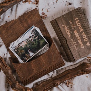 May include: A wooden photo box with a stack of printed photos inside. The box is open, revealing the photos. The lid of the box has the phrase "I LOVE YOU to the mountains and back" engraved on it. The box is surrounded by dried branches.