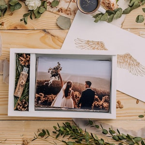 May include: A white wooden photo box with a wedding photo, a wooden USB drive, and pine cones. The box is on a wooden surface with greenery, a camera, and stationery with gold wing illustrations.