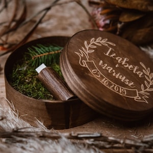 Op de afbeelding: Een ronde houten doos met een deksel, met daarin een USB-stick en mos. Het deksel is gegraveerd met de namen "Izabela i Mariusz" en de datum "05.10.2019" in een lauwerkransontwerp. De doos is open en onthult de inhoud.