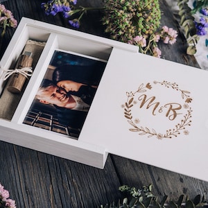 Può includere: Una scatola fotografica in legno bianco con coperchio scorrevole, contenente una fotografia, una chiavetta USB in legno e un sacchetto di lino. Il coperchio presenta una ghirlanda floreale con le iniziali "M&B".