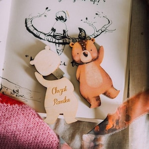 May include: Two wooden bear-shaped ornaments, one light-colored and one brown, are displayed on a book page with a whimsical illustration. The light-colored bear has text that reads "Chrzest Ramika". The brown bear has a spotted pattern.