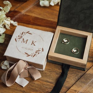Op de afbeelding: Een houten ringendoos met twee gouden ringen met hartdetails, geplaatst op een groen fluwelen interieur. Een bijpassende doos met de initialen "MK" en de datum "07.02.2025" staat ernaast. Beige lint en witte bloemen zijn ook aanwezig.
