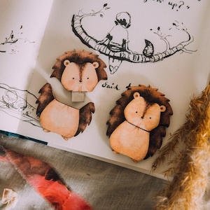 May include: Three wooden hedgehog-shaped USB flash drives are displayed on an open book with a drawing of a person on a branch. The hedgehogs have brown fur and tan faces. The book has handwritten text and illustrations.