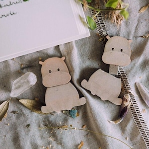 May include: Two wooden hippo-shaped paper clips with dark eyes and small ears. One clip is open, and the other is closed. The clips are light brown and sit on a gray fabric surface with dried leaves and flower petals.