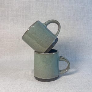May include: Two ceramic mugs in a stacked arrangement. The mugs are a muted teal color with a dark brown base and rim. Each mug has a rounded handle. The mugs are handmade and have a rustic, artisanal appearance.