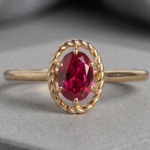 May include: A gold ring featuring a central oval-cut red gemstone held within a twisted rope-style metal frame. The band is smooth and polished, connecting to the decorative oval setting that surrounds the vibrant red stone in a secure prong mount.