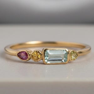 May include: A delicate gold ring featuring a rectangular light blue gemstone flanked by a pink, two yellow, and a light green gemstone. The ring is set on a white surface.
