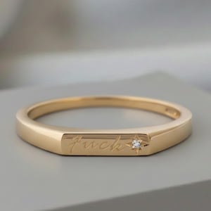 May include: A gold-colored ring with the word "fuck" engraved on the front. A small diamond is set next to the word. The ring is on a gray surface.