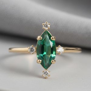 May include: A gold ring featuring a marquise-cut, emerald-green gemstone as the centerpiece. The ring is accented with small, round, clear gemstones on either side of the main stone, and on the band. The ring is set against a neutral background.