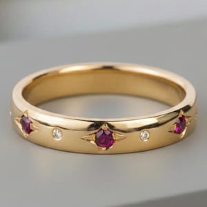 May include: A gold band ring featuring a series of alternating round red rubies and small clear white diamonds. The gemstones are set into the polished gold surface, with the rubies held in star-shaped settings and the diamonds in simple round bezels.