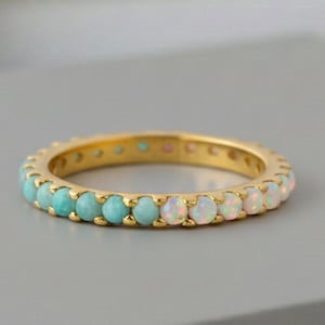 May include: A gold ring featuring a band of alternating turquoise and opal gemstones. The ring is displayed on a grey surface, highlighting the colours of the stones. The ring is a piece of jewellery.