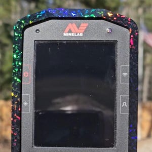 May include: A black and gray metal detector control panel with a screen and buttons. The device has a colorful, iridescent border. The brand name "Minelab" is visible at the top. The model name "Manticore" is printed on the device.