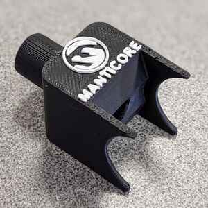 Op de afbeelding: Een zwart 3D-geprint object met het woord "MANTICORE" in witte letters. Het object heeft een cilindrisch uiteinde en een logo met een wit handontwerp. Het object heeft een getextureerd oppervlak en is waarschijnlijk een gereedschap of accessoire.