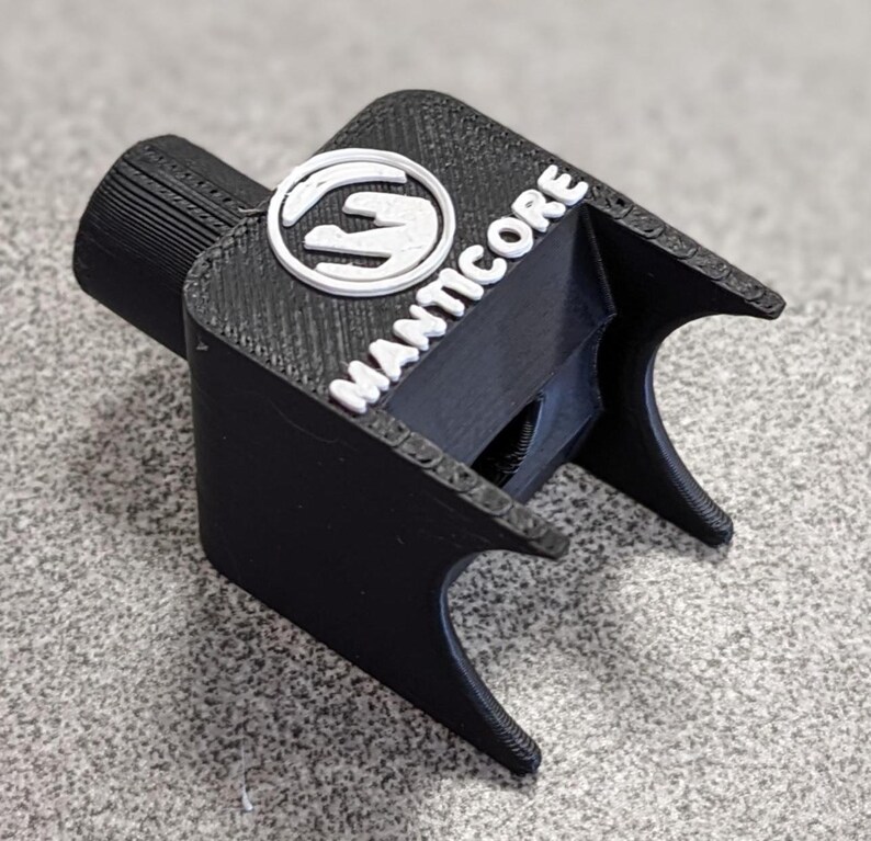 Op de afbeelding: Een zwart 3D-geprint object met het woord "MANTICORE" in witte letters. Het object heeft een cilindrisch uiteinde en een logo met een wit handontwerp. Het object heeft een getextureerd oppervlak en is waarschijnlijk een gereedschap of accessoire.