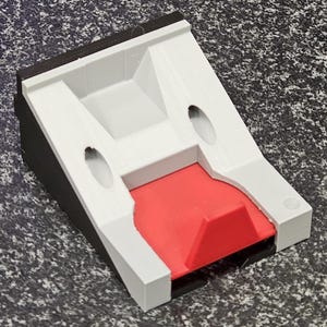 Op de afbeelding: Een 3D-geprint object met een wit, grijs, zwart en rood kleurenschema. Het object heeft een rechthoekige basis met een schuin ontwerp, twee ovale uitsparingen en een centraal verzonken gebied. Het rode gedeelte is een verhoogde, schuine vorm.