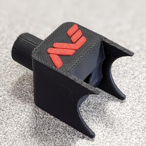Op de afbeelding: Een zwart 3D-geprint object met een rood logo. Het object heeft een cilindrisch deel en een U-vormig ontwerp. Het rode logo bestaat uit drie gestapelde, schuine lijnen. Het is waarschijnlijk een gereedschap of accessoire.