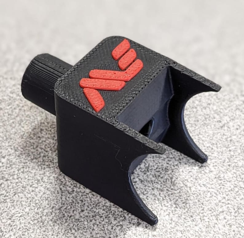 Op de afbeelding: Een zwart 3D-geprint object met een rood logo. Het object heeft een cilindrisch deel en een U-vormig ontwerp. Het rode logo bestaat uit drie gestapelde, schuine lijnen. Het is waarschijnlijk een gereedschap of accessoire.