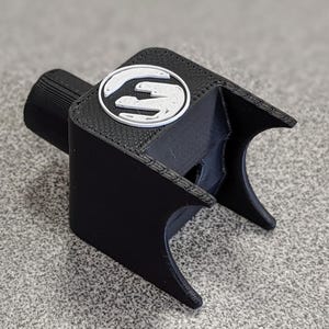 Op de afbeelding: Een zwart 3D-geprint object met een wit en grijs logo. Het object heeft een cilindrisch stuk aan &eacute;&eacute;n uiteinde en een gebogen, open ontwerp aan de andere kant. Het logo is een gestileerde "S" in een cirkel.