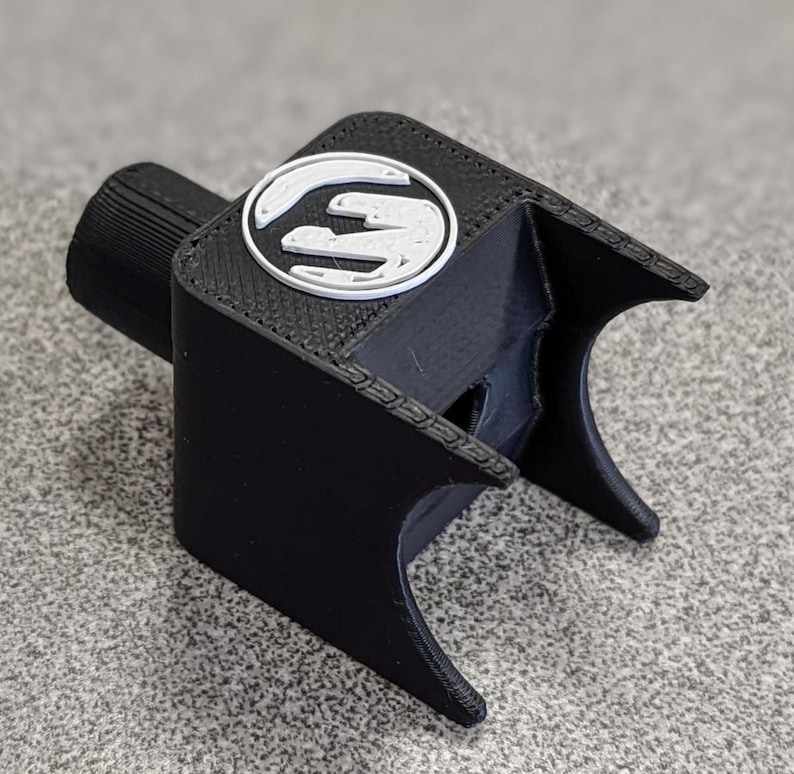 Op de afbeelding: Een zwart 3D-geprint object met een wit en grijs logo. Het object heeft een cilindrisch stuk aan &eacute;&eacute;n uiteinde en een gebogen, open ontwerp aan de andere kant. Het logo is een gestileerde "S" in een cirkel.