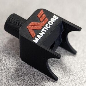 Op de afbeelding: Een zwart 3D-geprint object met het woord "MANTICORE" in wit en een rood logo. Het object heeft een cilindrisch gedeelte en een rechthoekige basis met gebogen randen. Het is waarschijnlijk een onderdeel of accessoire.