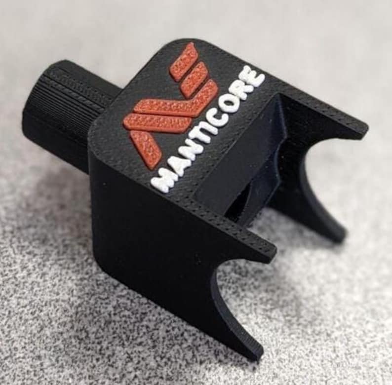 Op de afbeelding: Een zwart 3D-geprint object met het woord "MANTICORE" in wit en een rood logo. Het object heeft een cilindrisch gedeelte en een rechthoekige basis met gebogen randen. Het is waarschijnlijk een onderdeel of accessoire.