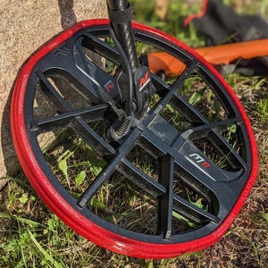 May include: A black metal detector coil with a vibrant red edge, resting on grass. The coil has a grid-like design and is connected to a black cable. The brand name is visible on the coil.